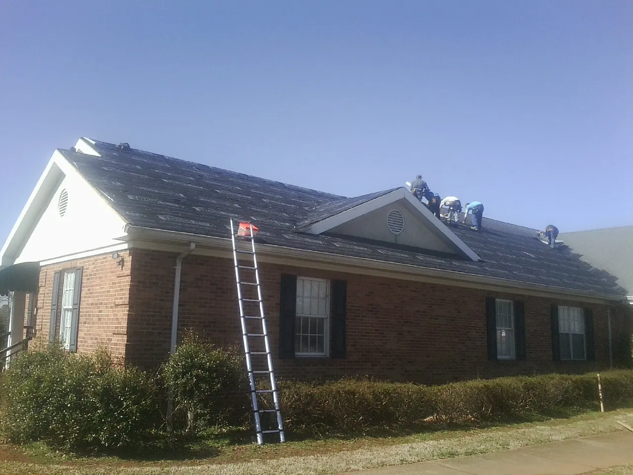 Crew tearing shingles off a brick ranch roof with a ladder set — Metal Roof Repair work by our crew in Saint Albans
